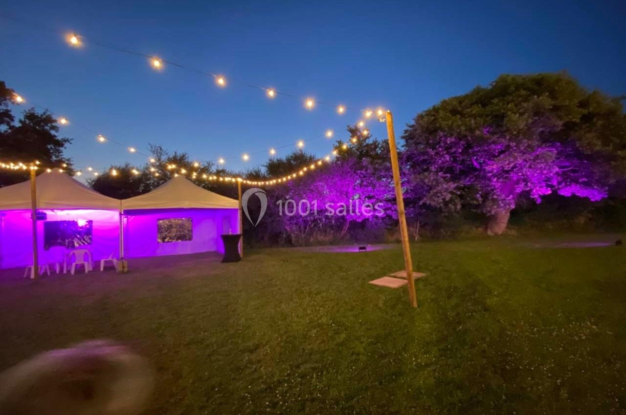 Tentes blanches éclairées par des guirlandes lumineuses et projecteurs violets dans un jardin au crépuscule.