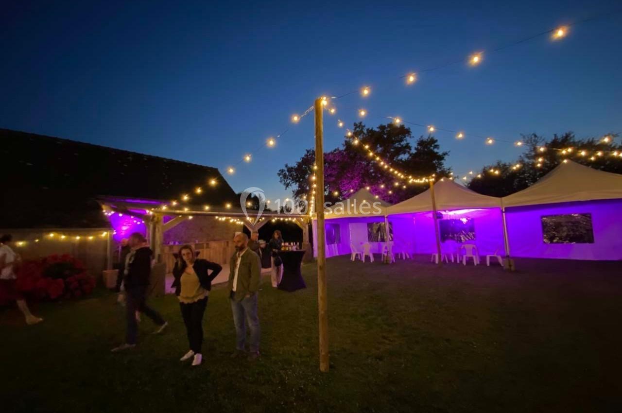 Tentes éclairées en violet et guirlandes lumineuses dans un jardin au crépuscule, avec quelques personnes présentes.