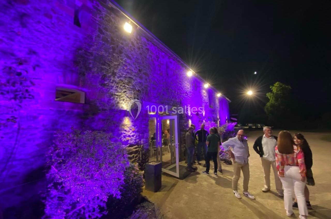 Façade en pierre éclairée de lumières violettes la nuit, avec des personnes discutant à l'extérieur.