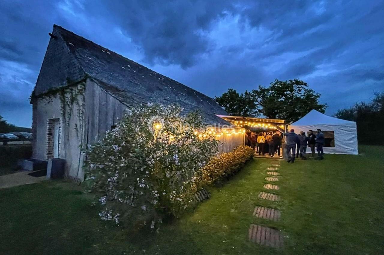 Bâtiment en pierre éclairé par des guirlandes lumineuses, avec des invités rassemblés sous un auvent au crépuscule.