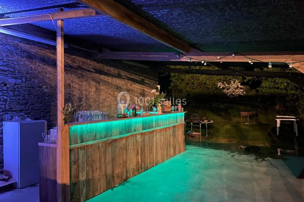 Bar en bois éclairé par des lumières LED vertes sous un abri extérieur, avec un jardin en arrière-plan.