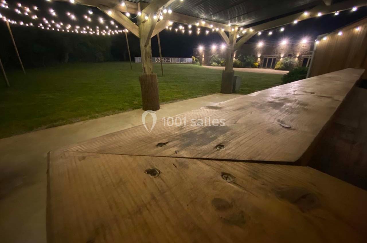 Terrasse en bois éclairée par des guirlandes lumineuses, avec vue sur une pelouse et des bâtiments en arrière-plan.
