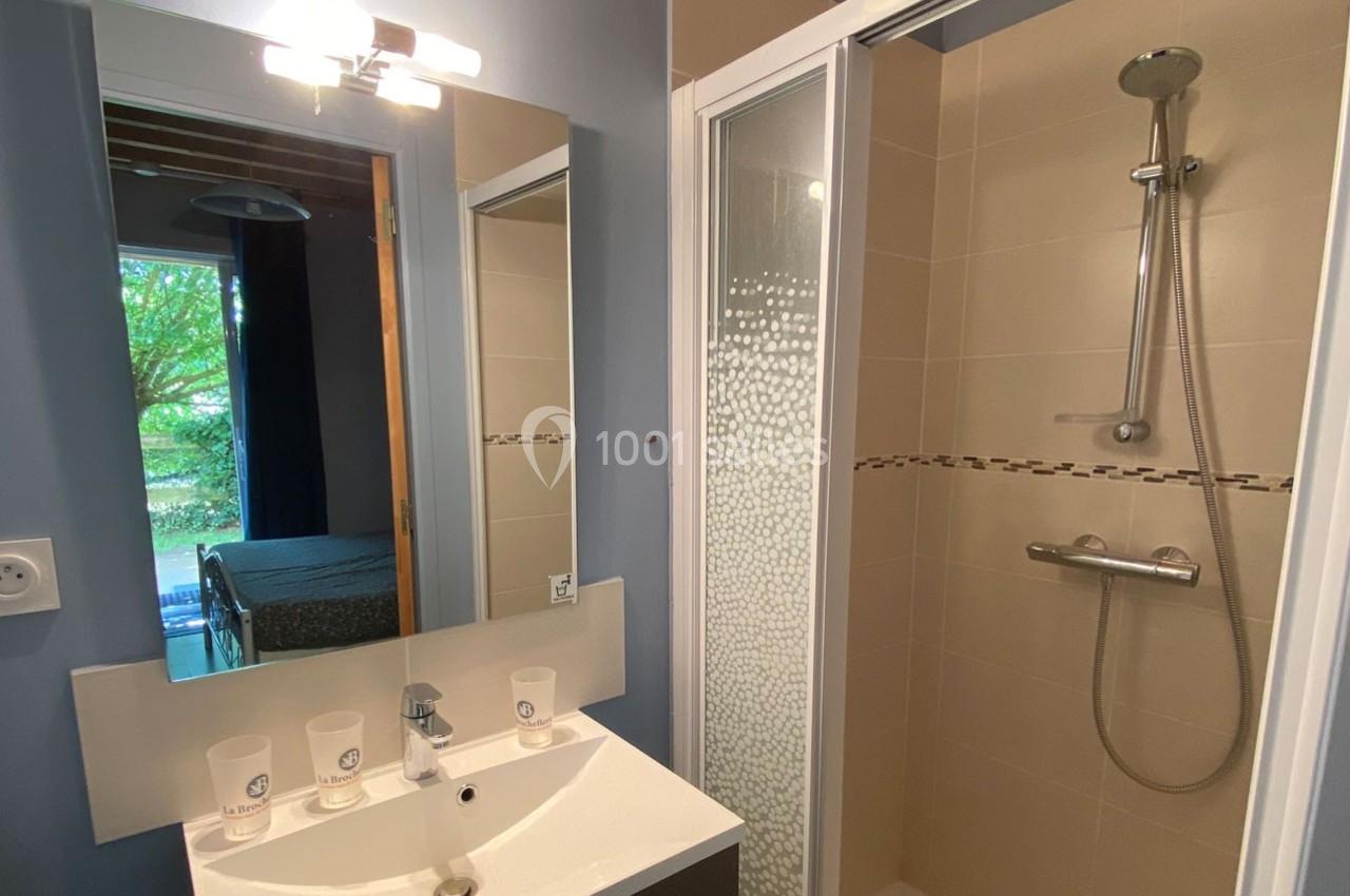Salle de bain avec lavabo, miroir éclairé, cabine de douche vitrée et vue partielle sur une chambre au fond.