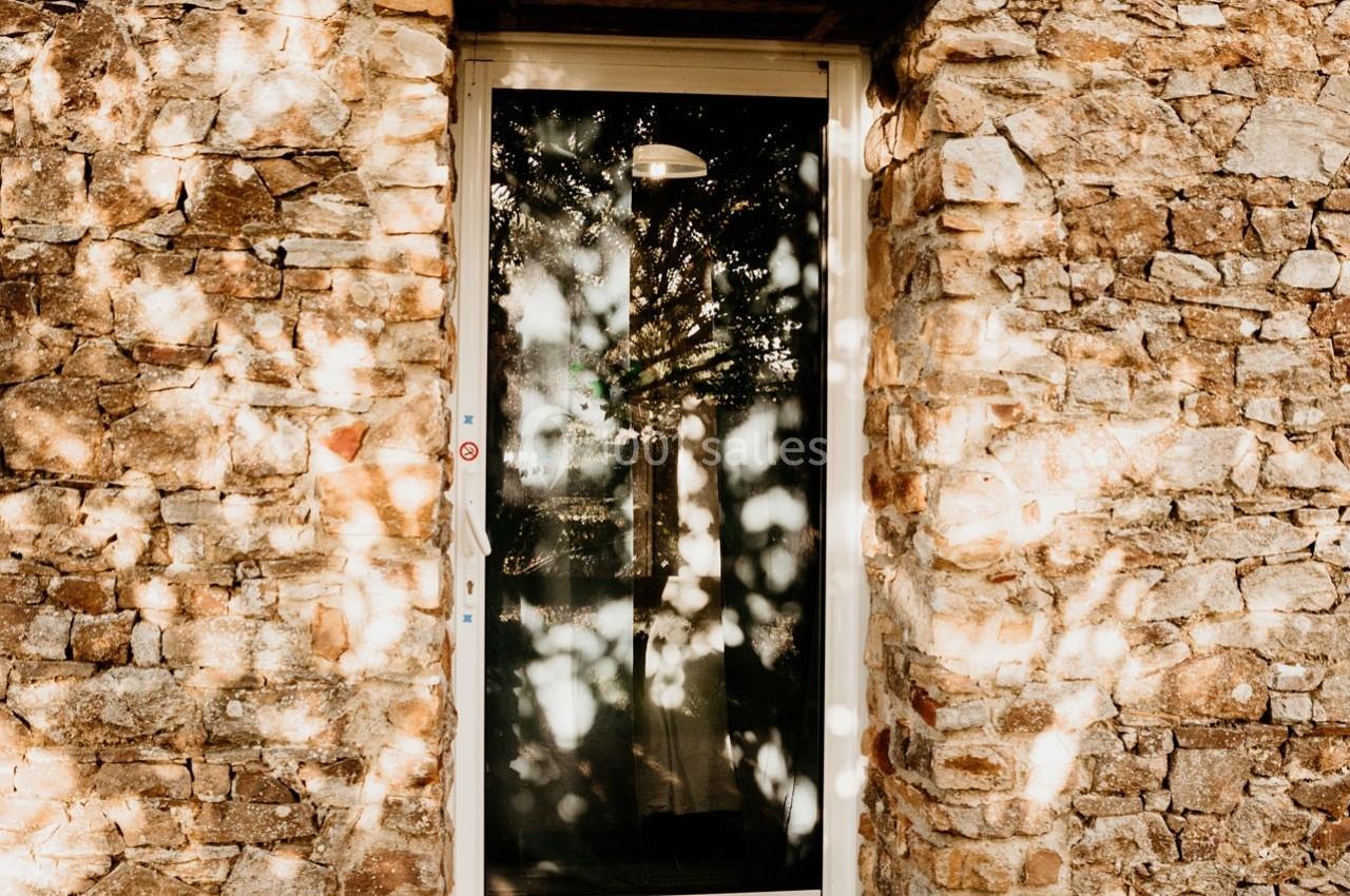 Porte vitrée encadrée par un mur en pierre, avec des ombres d'arbres projetées sur la surface.