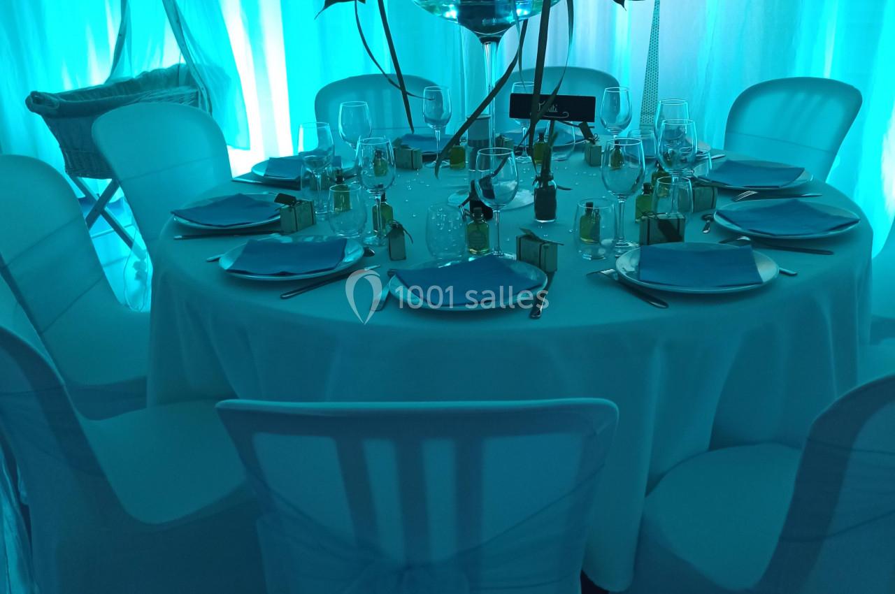 Table ronde décorée avec nappes blanches, vaisselle élégante et centre de table floral, éclairage bleu en arrière-plan.