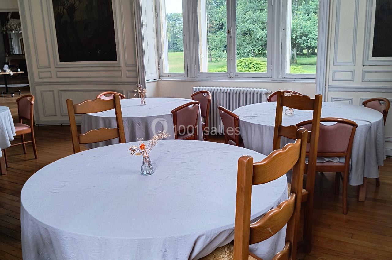 Salle lumineuse avec tables rondes couvertes de nappes blanches, chaises en bois et vue sur un jardin à travers de grandes…