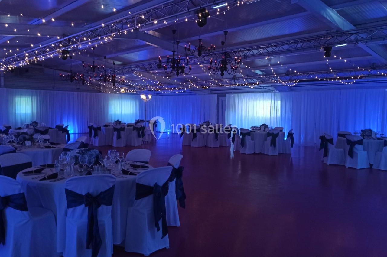 Salle de réception décorée avec des tables rondes, nappes blanches, chaises ornées de rubans et éclairage bleu tamisé.