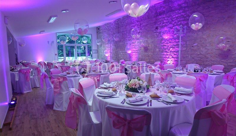 Salle de réception décorée avec des nappes blanches, des rubans roses et des ballons, éclairée par une lumière violette.
