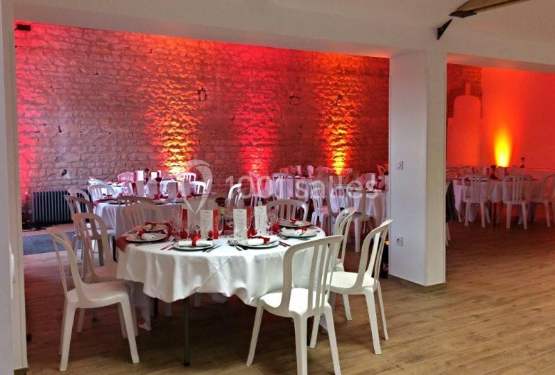 Salle de réception décorée avec des tables rondes dressées, des nappes blanches et un éclairage mural orange.