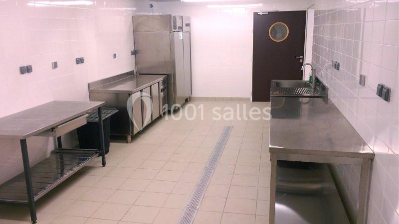 Cuisine professionnelle avec plans de travail en inox, réfrigérateurs, évier et sol carrelé beige.