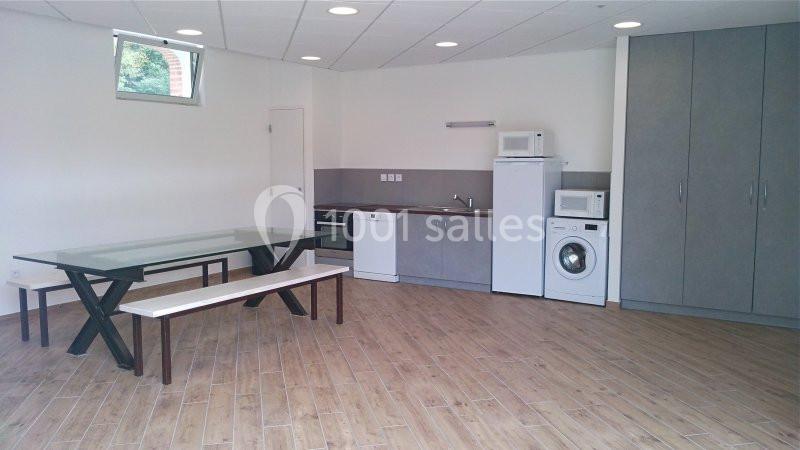 Salle équipée avec table, banc, kitchenette, lave-vaisselle, micro-ondes, lave-linge et placards sur un sol en parquet.