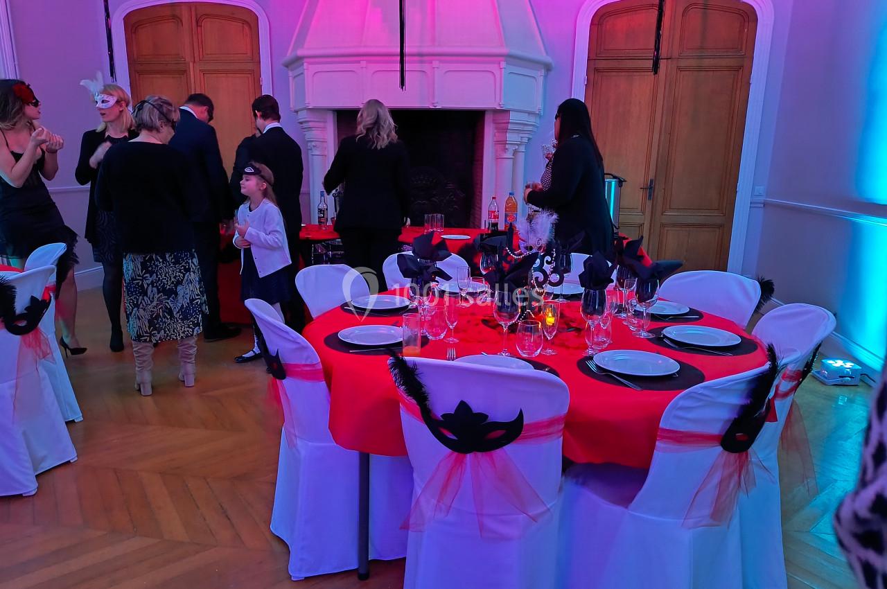 Salle décorée pour un événement, avec tables dressées en rouge et blanc, ornées de masques noirs et plumes.