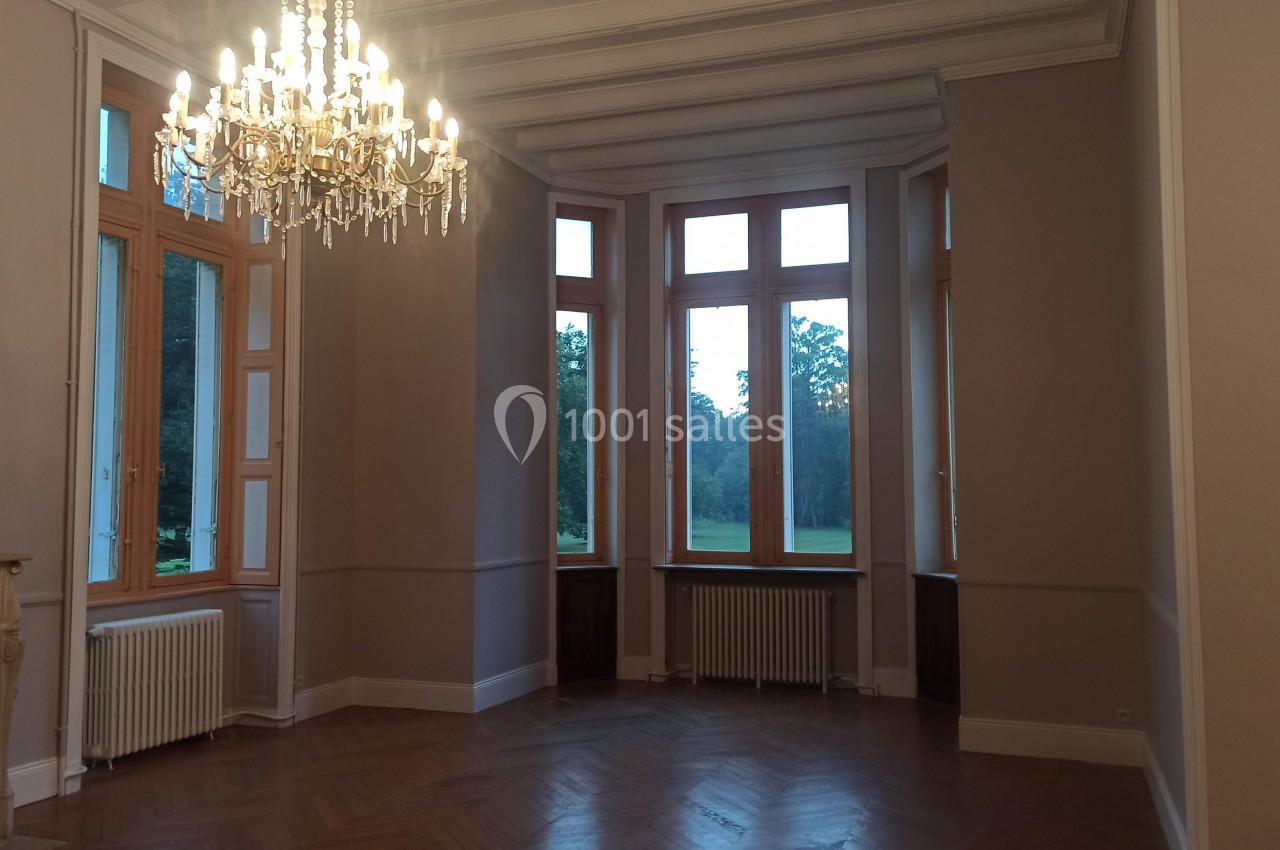 Salle lumineuse avec parquet en bois, grandes fenêtres donnant sur un parc, et un lustre suspendu au plafond.