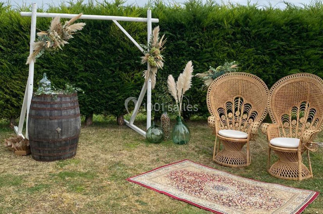 Décoration extérieure avec fauteuils en rotin, tapis oriental, tonneau en bois et ornements végétaux devant une haie.