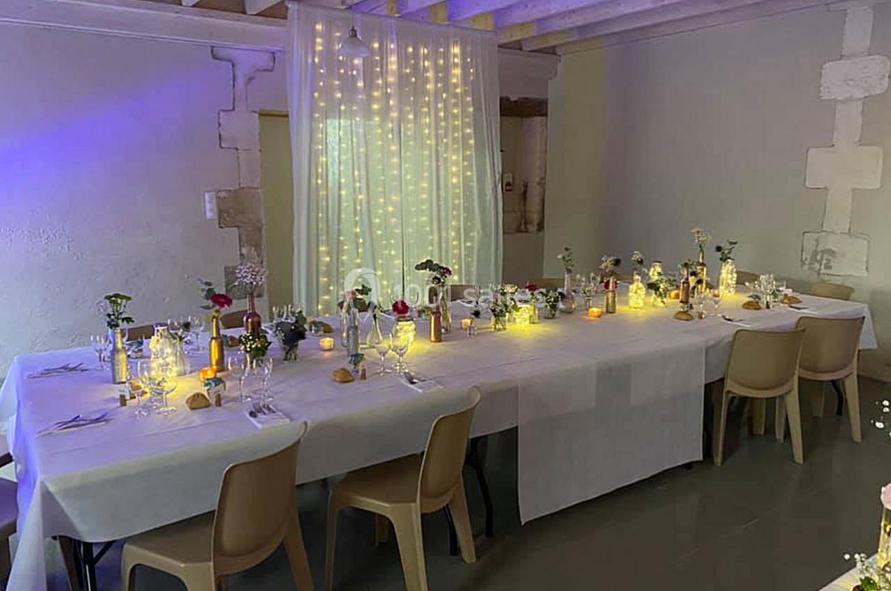 Grande table décorée pour un repas, ornée de fleurs, bougies et guirlandes lumineuses dans une salle au plafond en bois.