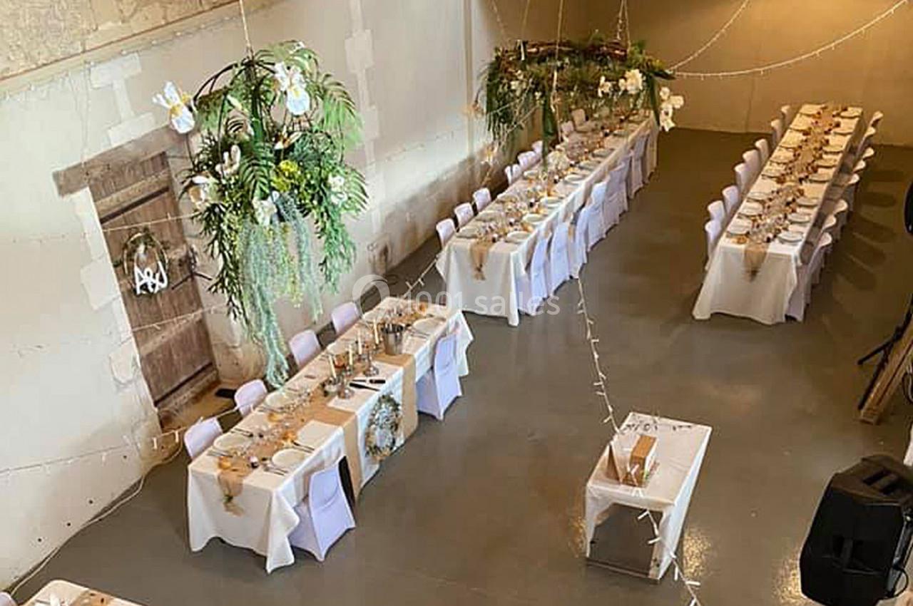 Salle de réception décorée pour un événement, avec longues tables dressées, nappes blanches et ornements floraux suspendus.