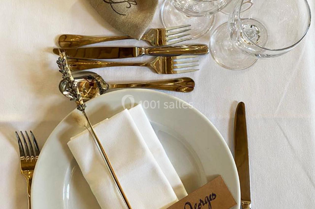 Table dressée avec assiette blanche, serviette pliée, couverts dorés, verres et étiquette portant un prénom.