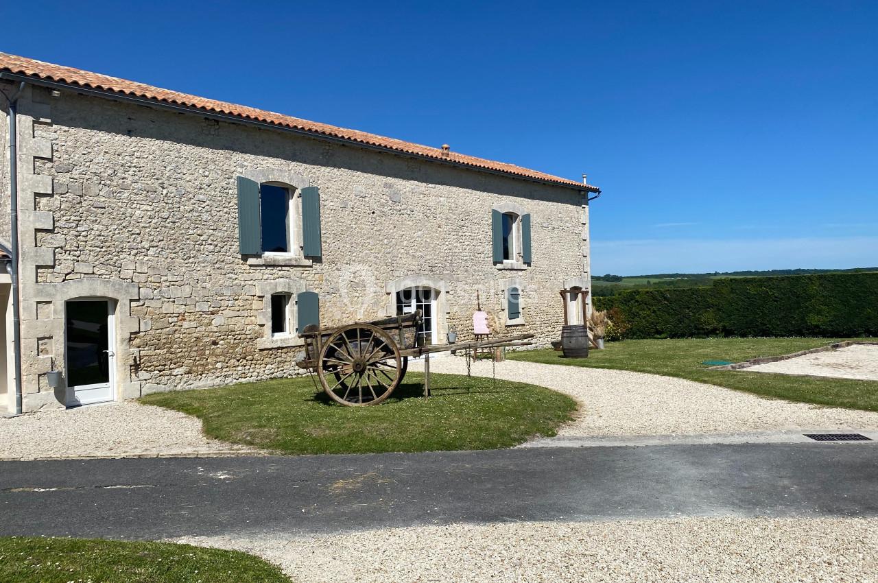 Façade en pierre d'une maison traditionnelle avec volets bleus, une charrette en bois et un jardin ensoleillé.