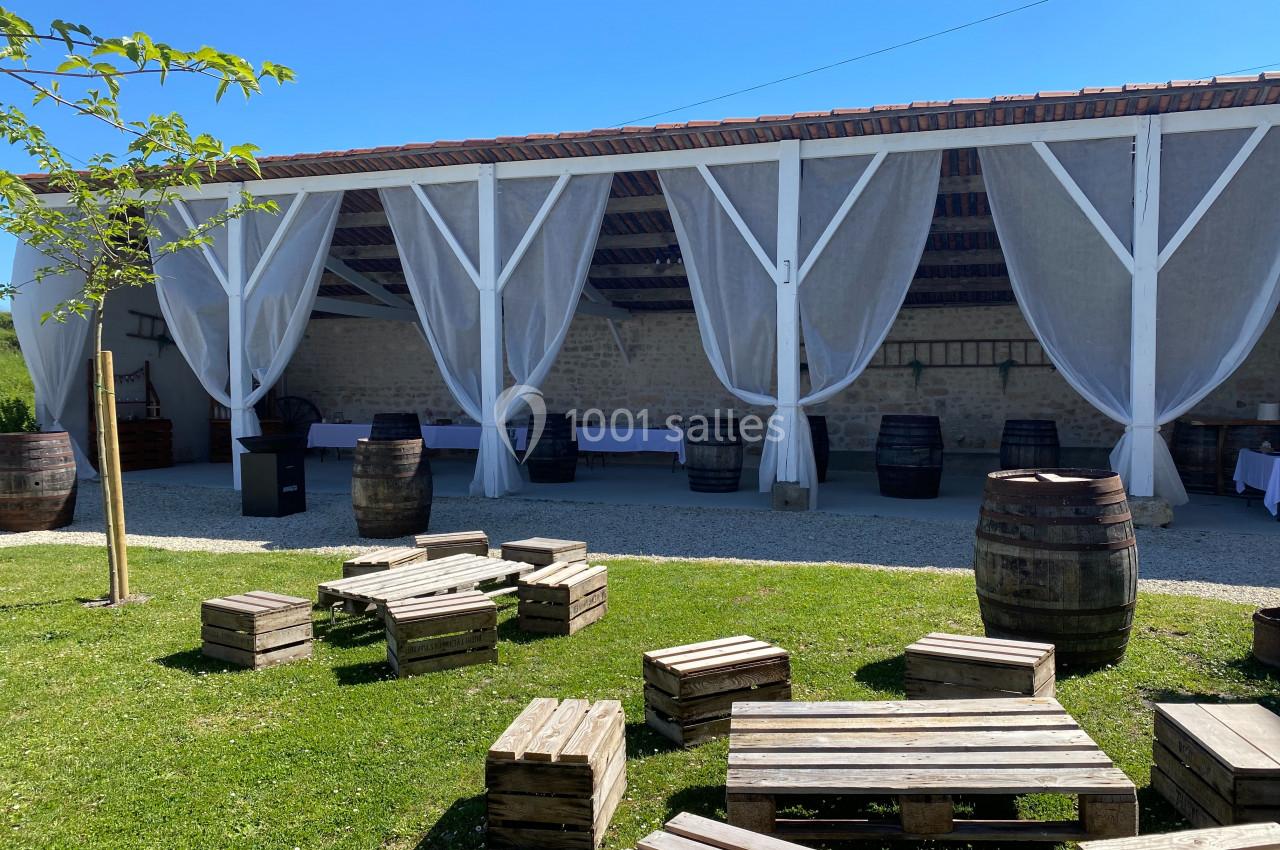 Terrasse couverte avec rideaux blancs, tonneaux en bois et palettes disposées sur une pelouse par une journée ensoleillée.
