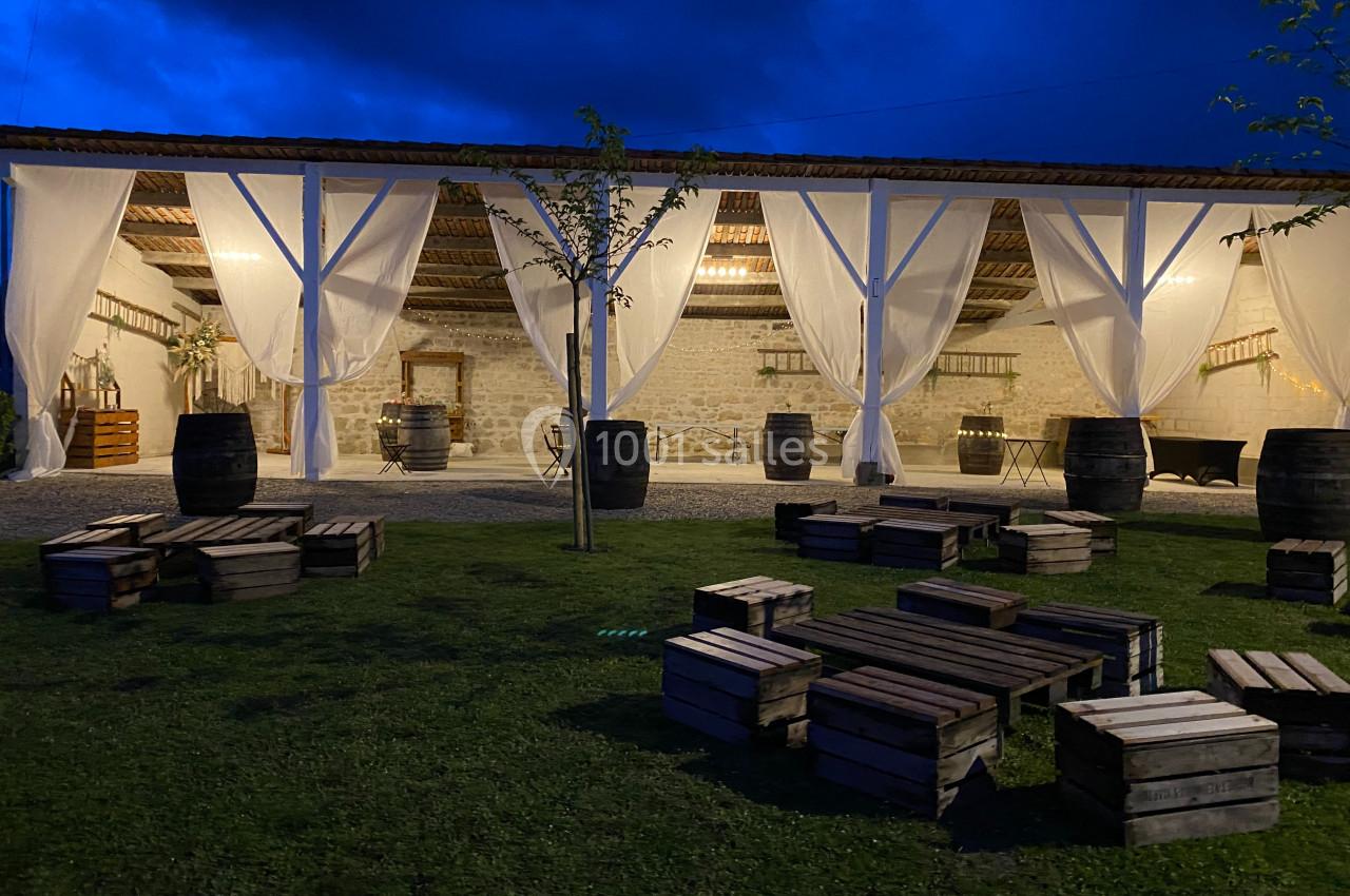 Structure en bois ouverte éclairée le soir, avec rideaux blancs, tonneaux et palettes disposés sur une pelouse.