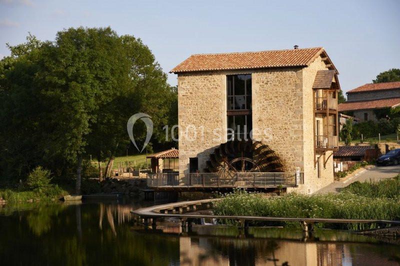 Bâtiment en pierre avec une roue à aubes, situé au bord d'un plan d'eau entouré de végétation.