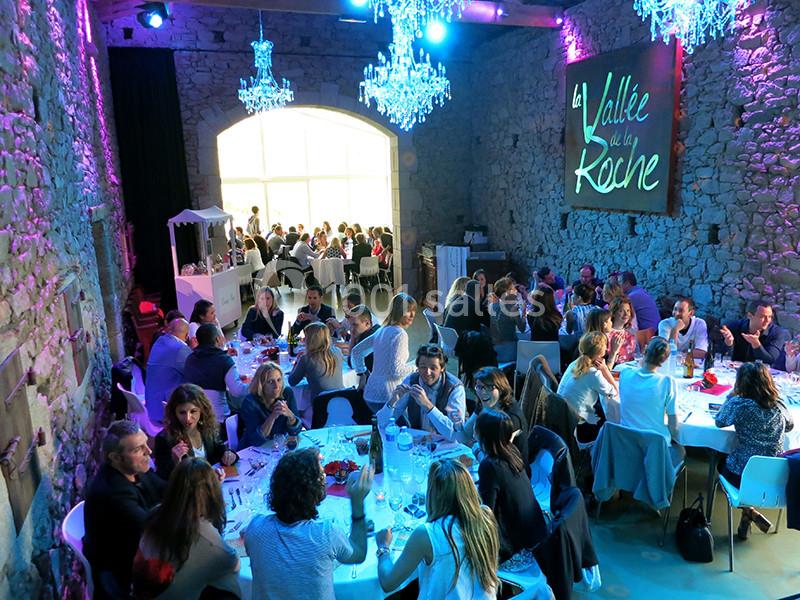 Salle en pierre avec des convives assis à des tables rondes, éclairée par des lustres et une lumière colorée.