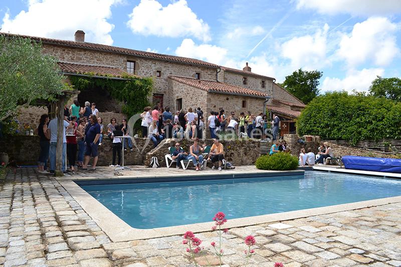 Groupe de personnes rassemblées près d'une piscine dans la cour d'une grande maison en pierre sous un ciel ensoleillé.