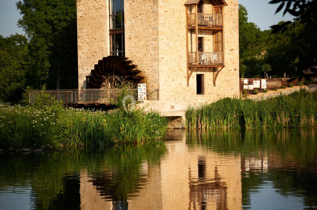 Bâtiment en pierre avec une roue à aubes, bordé par un plan d'eau et entouré de végétation.