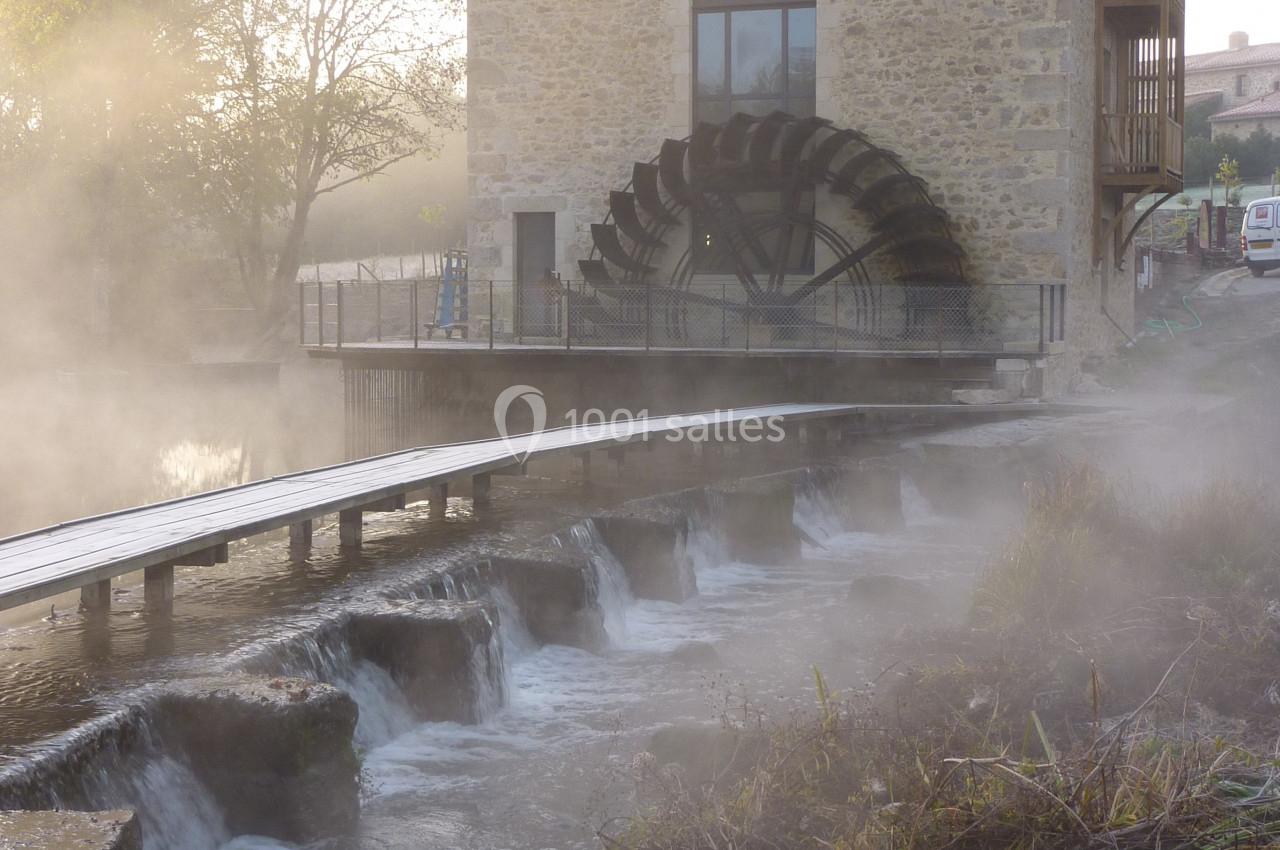 Bâtiment en pierre avec une grande roue à aubes, bordé par une rivière et une passerelle en bois dans une ambiance brumeuse.