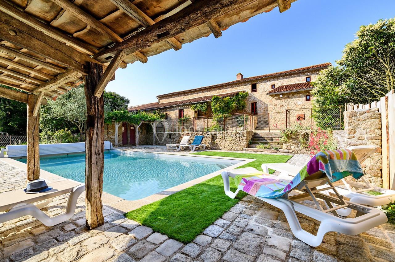 Piscine extérieure entourée de chaises longues, pelouse et terrasse en pierre, devant une maison en pierre.