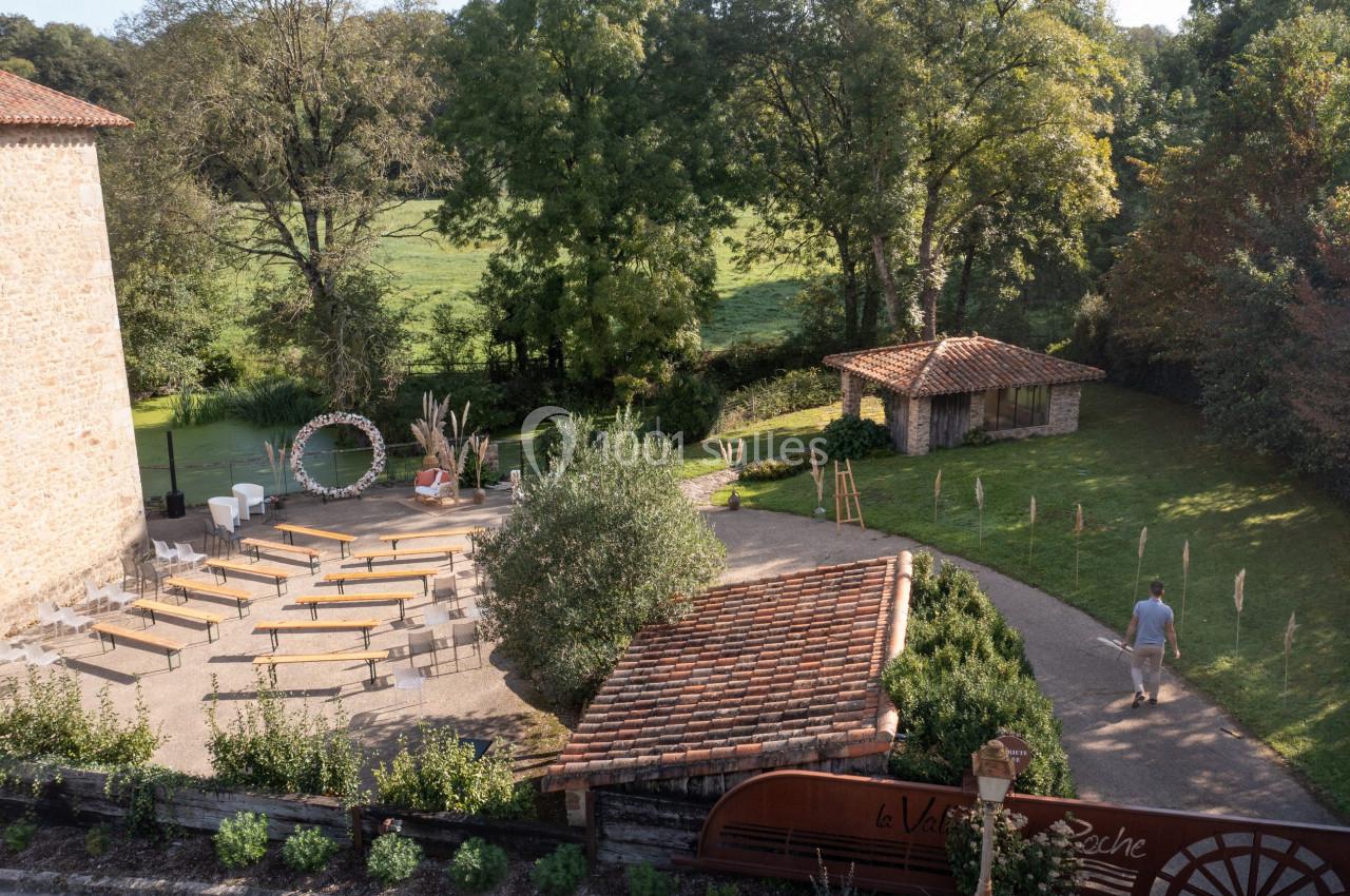 Vue d'un espace extérieur aménagé pour une cérémonie, avec des bancs, une arche florale et un jardin verdoyant.