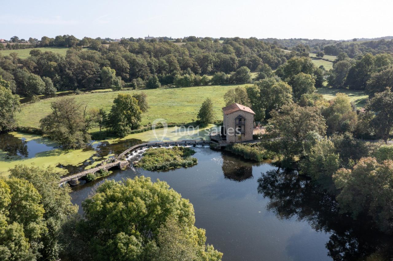 Vue aérienne d'un moulin en pierre près d'une rivière entourée de verdure et de paysages boisés.