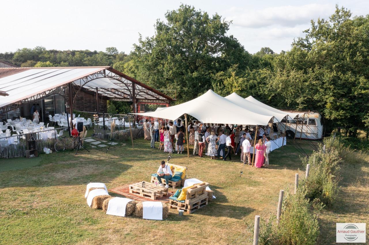 Vue d'une réception en plein air avec des invités sous une tente, des tables décorées et des bottes de foin en assises.