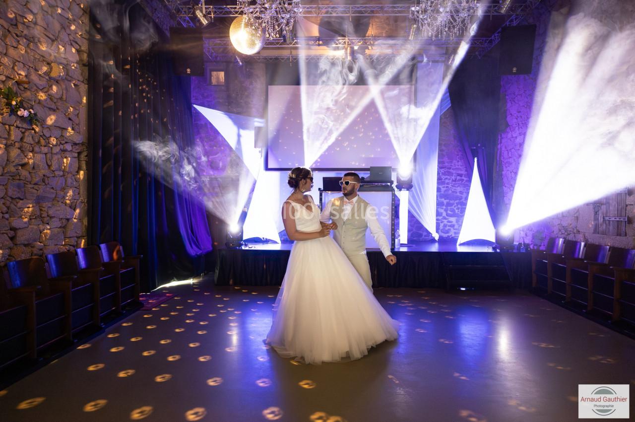 Un couple en tenue de mariage danse sur une piste éclairée par des jeux de lumière dans une salle en pierre.