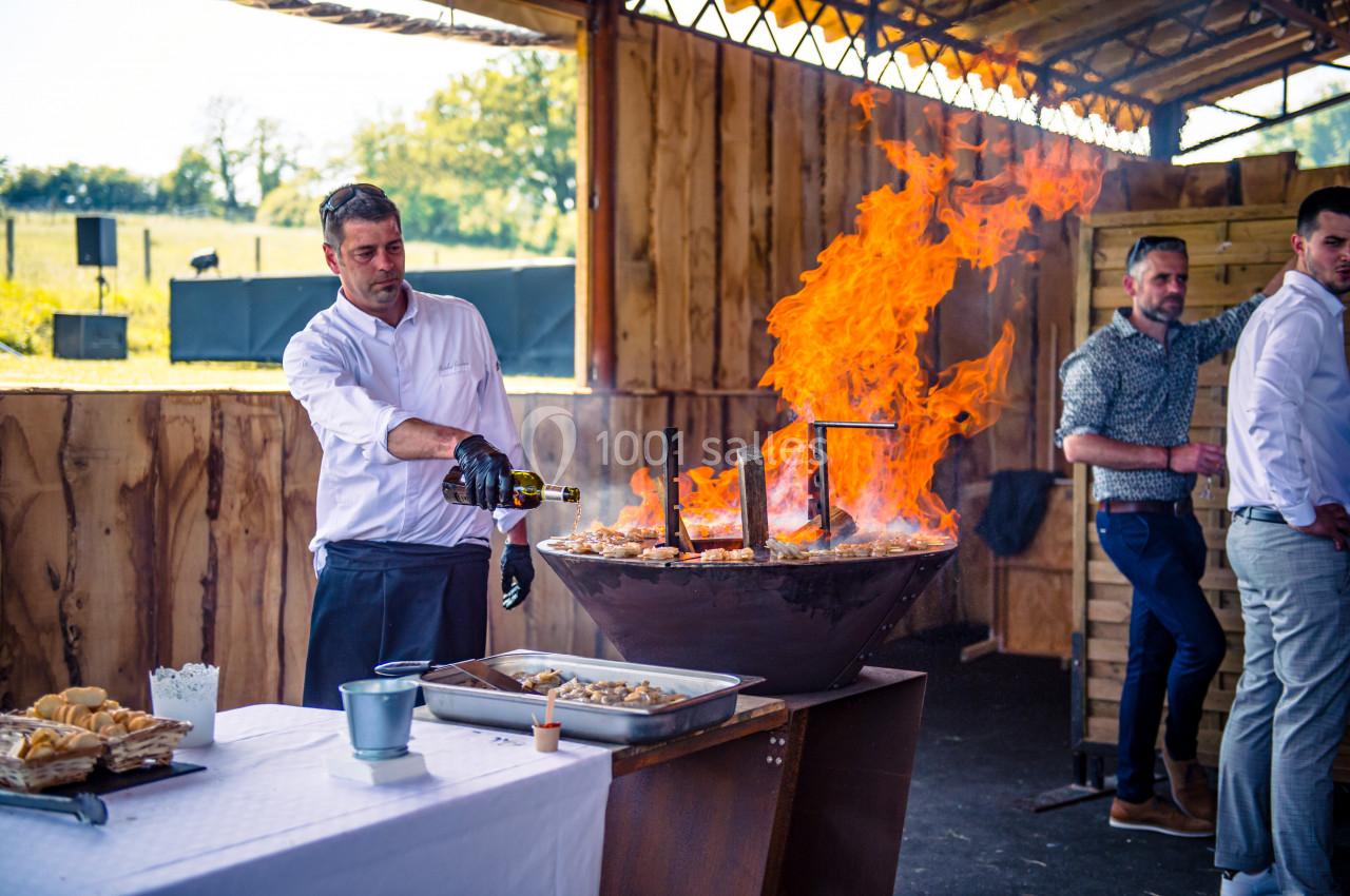 Un chef verse du liquide sur une grande plancha enflammée lors d'une cuisson en extérieur sous un abri en bois.