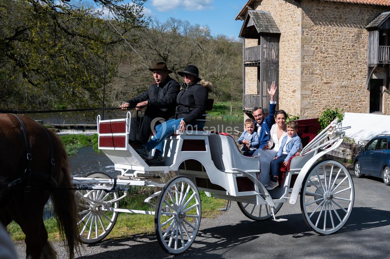 Un groupe de personnes en habits élégants est assis dans une calèche tirée par un cheval, près d'un bâtiment en pierre.