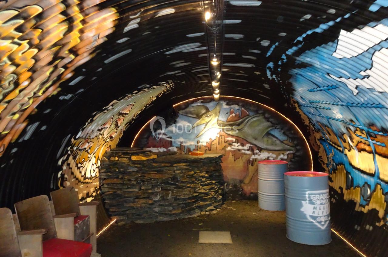 Intérieur d'un abri souterrain décoré de fresques colorées, avec des sièges en bois et des barils métalliques.
