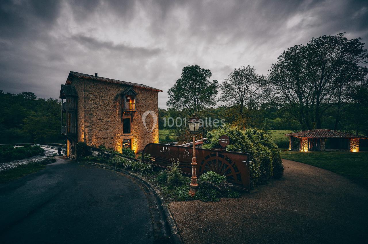 Bâtiment en pierre avec une roue à aubes, entouré de verdure, sous un ciel nuageux en fin de journée.