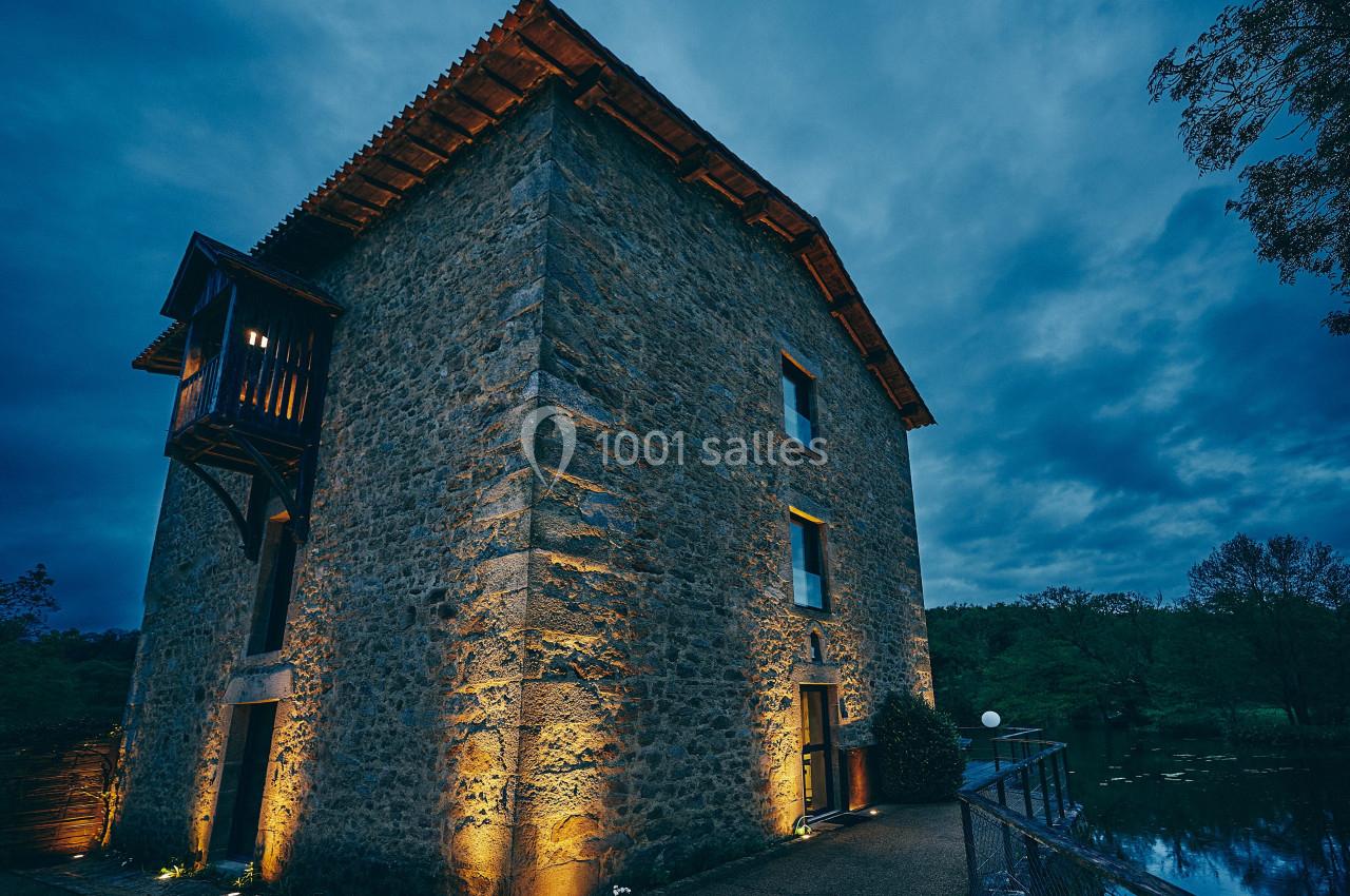 Bâtiment en pierre éclairé la nuit, avec un balcon en bois, situé près d'un plan d'eau et entouré de végétation.