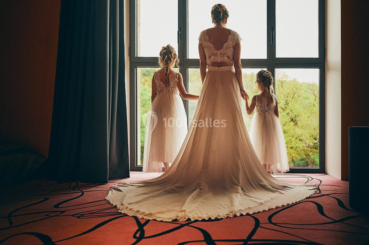 Une mariée en robe blanche tient la main de deux enfants devant une grande fenêtre donnant sur un paysage verdoyant.