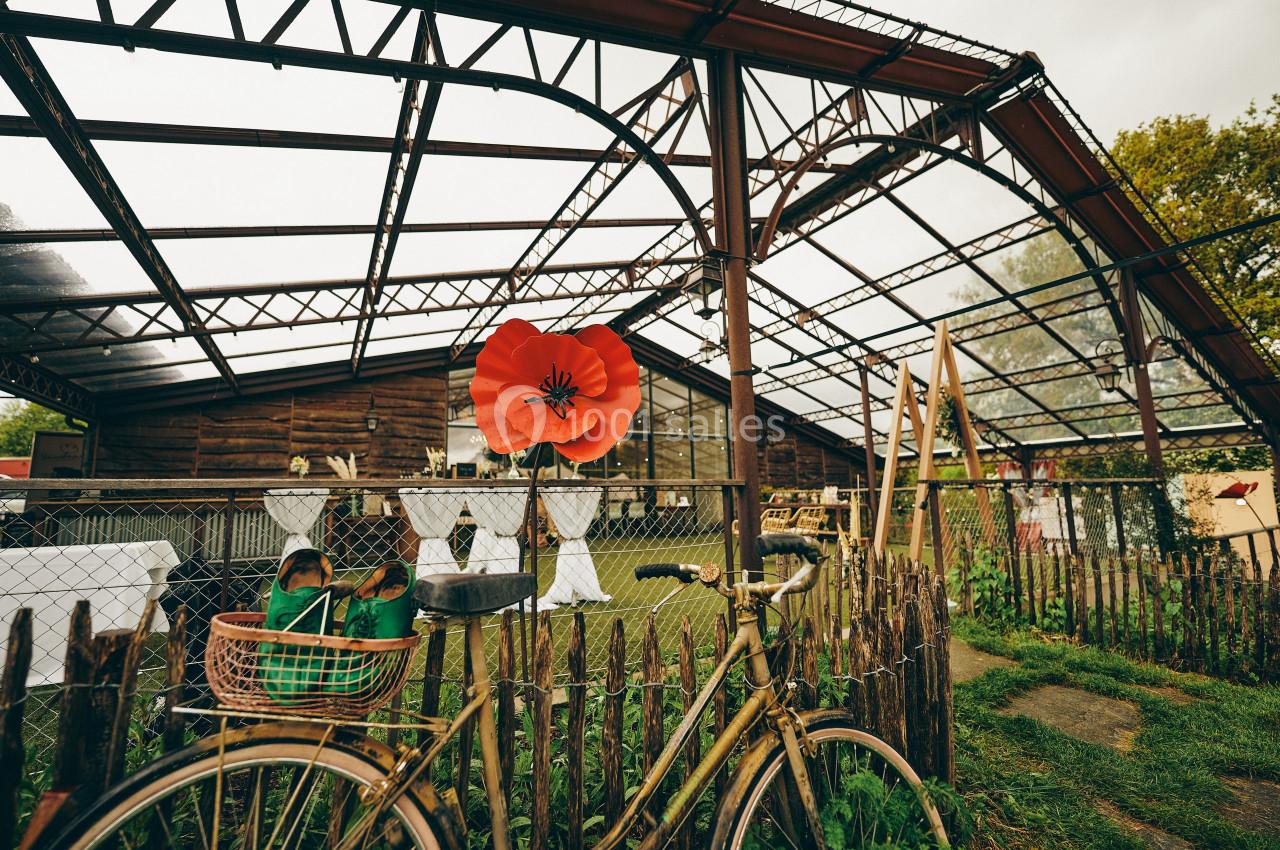 Vélo ancien appuyé contre une clôture en bois devant une serre en métal et verre partiellement ouverte.