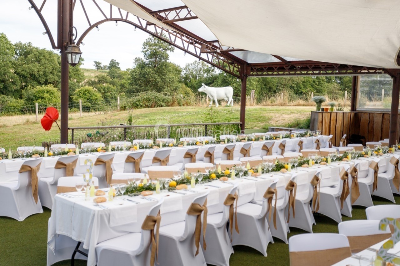 Tables de banquet décorées avec nappes blanches et rubans dorés, disposées en extérieur sous une structure couverte.