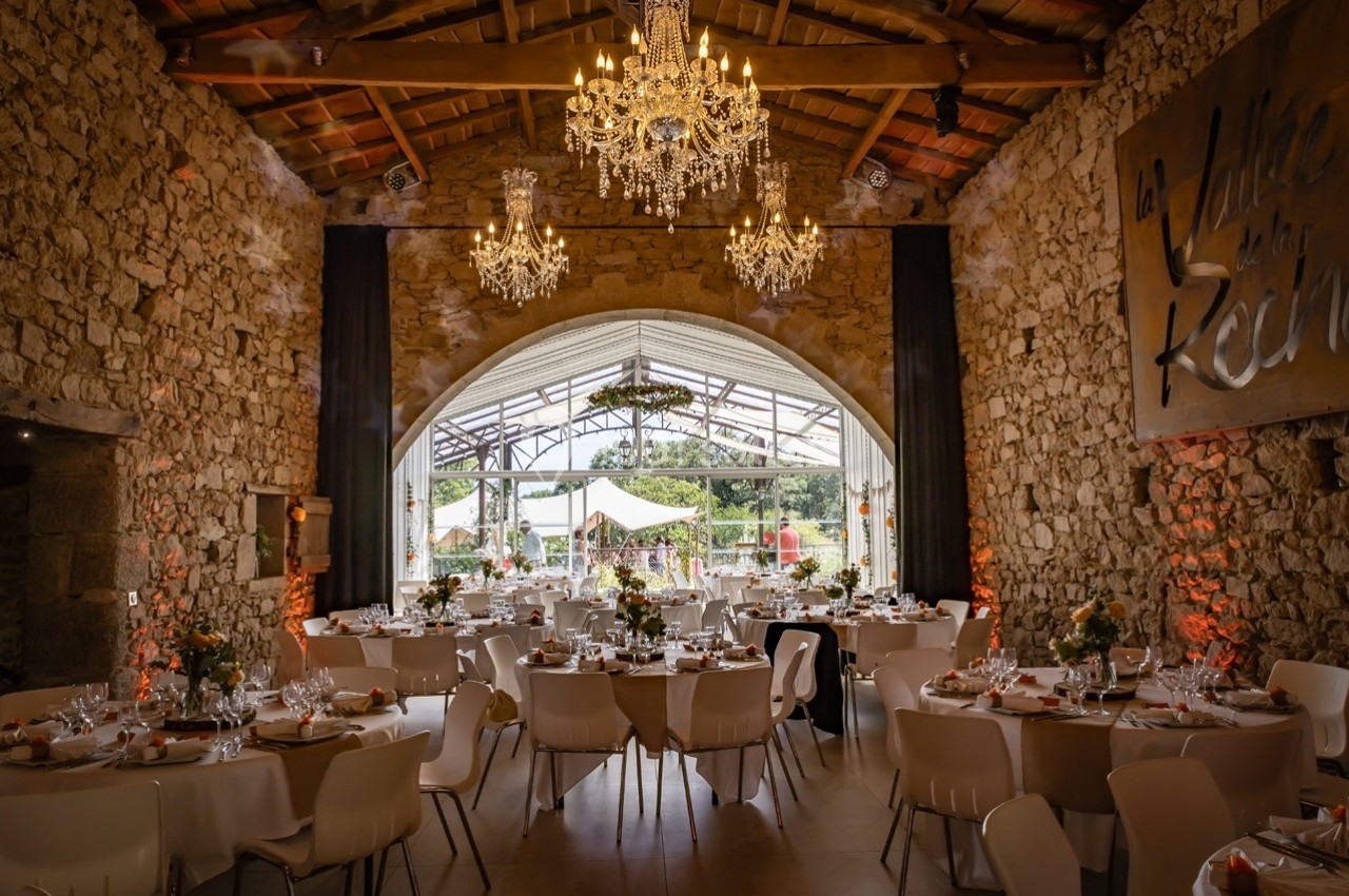 Salle de réception en pierre avec tables décorées, lustres suspendus et grande baie vitrée donnant sur l'extérieur.