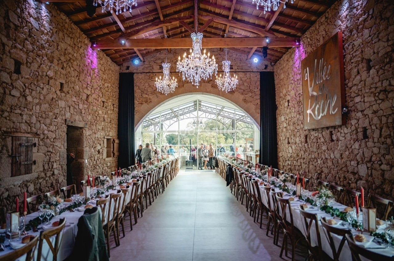 Salle de réception en pierre avec tables décorées, lustres suspendus et vue sur un jardin à travers une verrière.