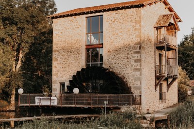 Ancien moulin en pierre avec une grande roue à aubes, bordé par un cours d'eau et entouré de végétation.