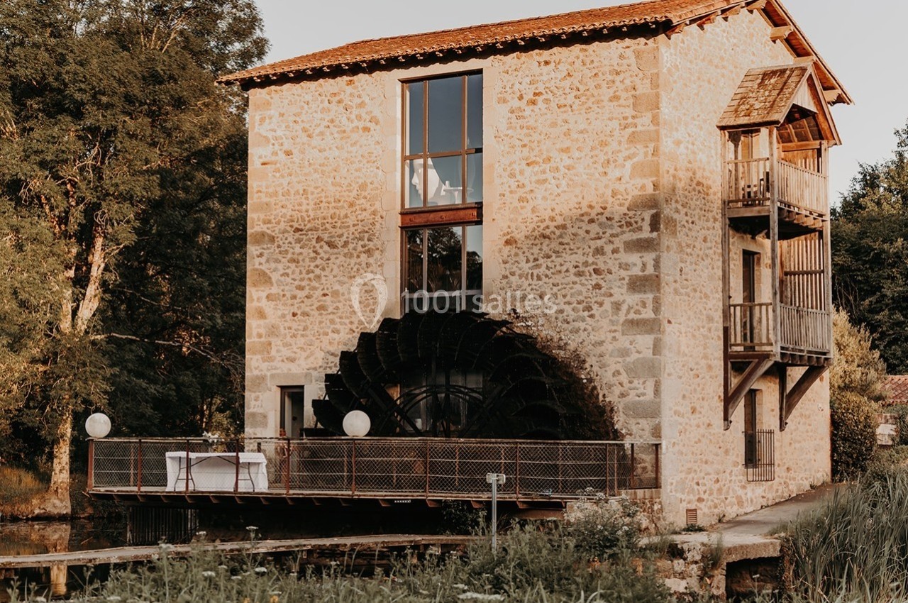 Ancien moulin en pierre avec une grande roue à aubes, bordé par un cours d'eau et entouré de végétation.