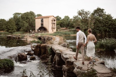 Ancien moulin en pierre avec une grande roue à aubes, bordé par un cours d'eau et entouré de végétation.