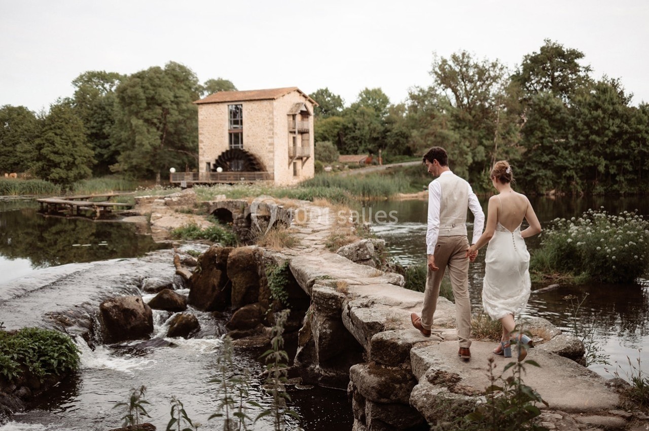 Un couple marche sur un chemin de pierres traversant une rivière, avec un moulin en pierre en arrière-plan.