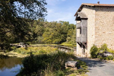 Ancien moulin en pierre avec une grande roue à aubes, bordé par un cours d'eau et entouré de végétation.