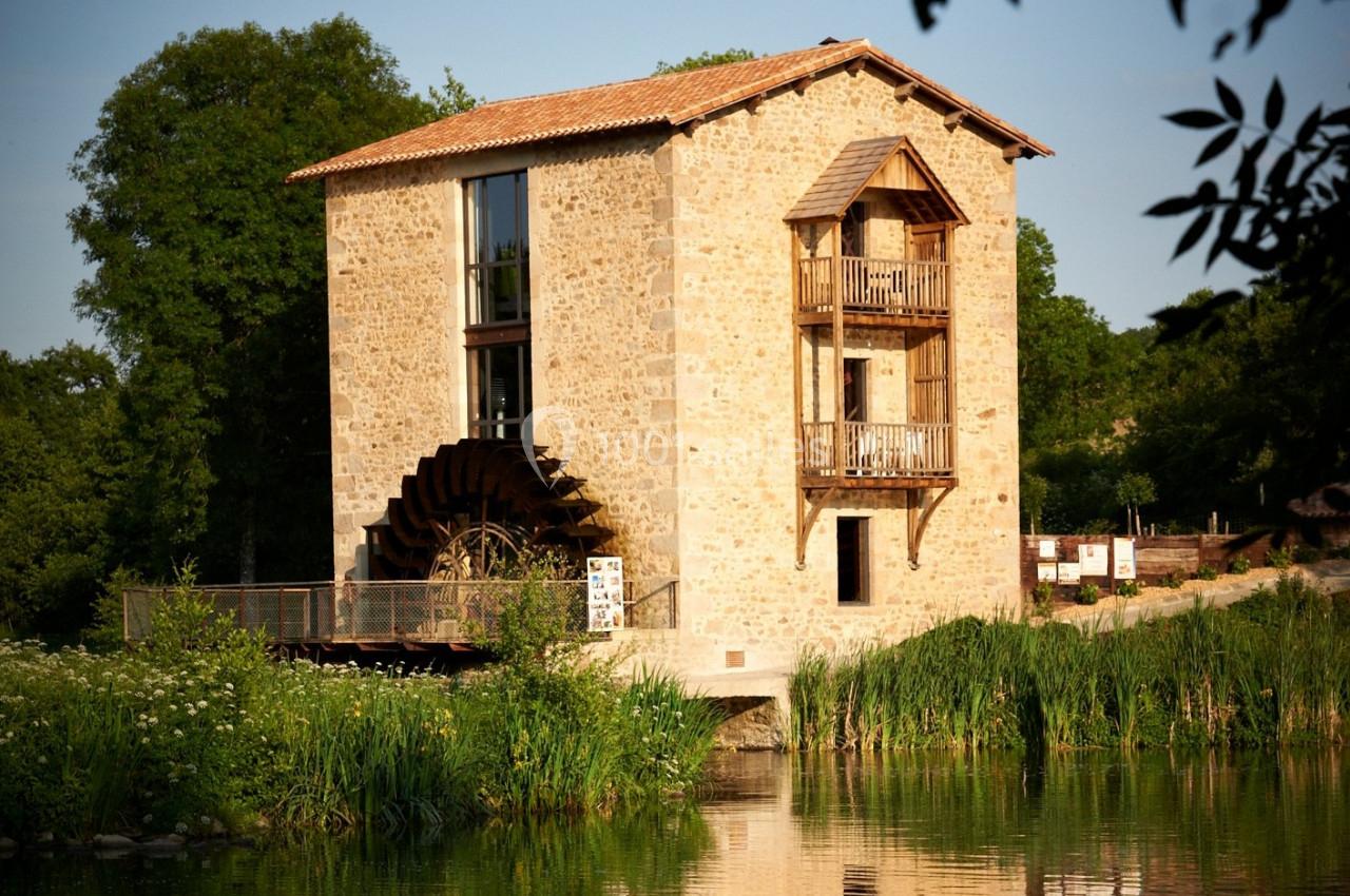 Moulin en pierre avec roue à aubes, entouré de végétation et bordant un plan d'eau calme.